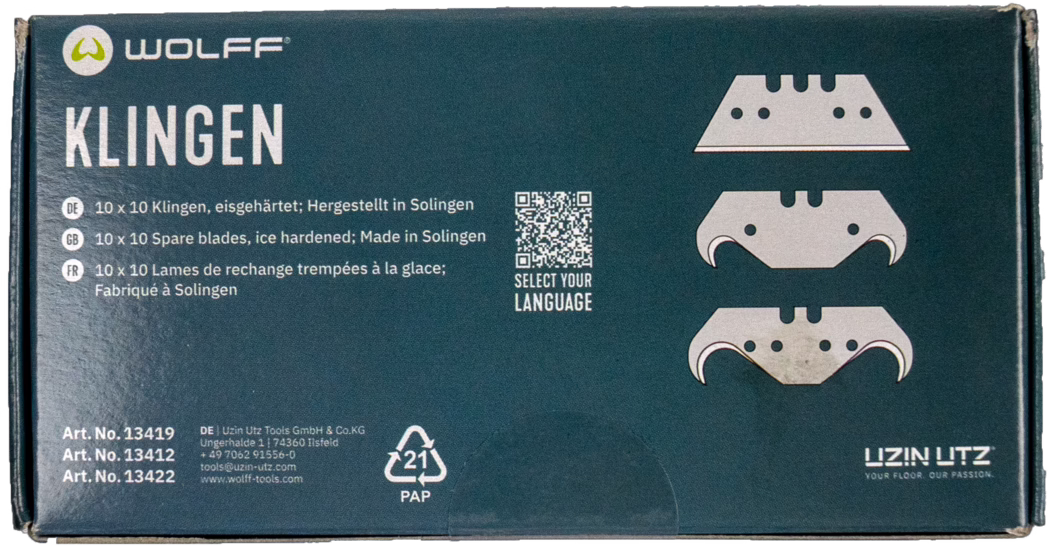 WOLFF_Dummy_13419_13421_blade box_2025-08 Close-up photograph of a rectangular cardboard box of Wolff replacement blades for floor tools. The dark teal packaging displays the brand logo 'WOLFF' at the top left and the large white word 'KLINGEN' on the left side. Three stylized blade illustrations are printed on the right; a small QR code with the caption 'SELECT YOUR LANGUAGE' sits between text blocks. Multilingual product lines name '10 x 10 spare blades, ice hardened; Made in Solingen' in German, English and French. Bottom-left shows three article numbers and manufacturer contact details, and a '21 PAP' recycling symbol appears near the lower center. The box edges show slight wear and handling marks. Overall a clean product package photographed on a white background.